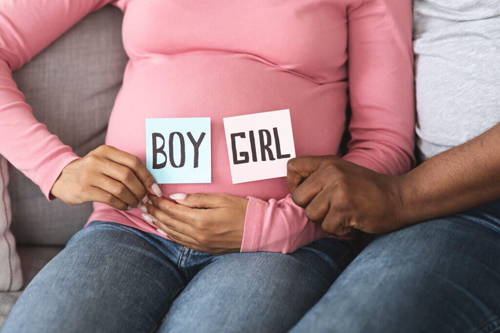 Gender reveal concept. Unrecognizable black pregnant couple holding pink and blue color card for baby boy or girl, closeup. African husband and wife guessing baby gender, sitting on couch at home